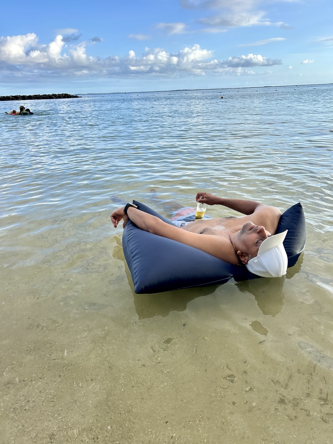 Rocky reclining on a dark blue float in clear shallow ocean water, wearing a white visor and holding a drink